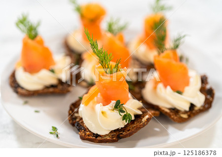 A gourmet smoked salmon canape elegantly arranged on a crispy cracker, topped with a swirl of smooth cream cheese, fresh thyme, and a drizzle of olive oil. The vibrant orange salmon contrasts 125186378