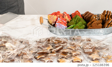 Captured from a mid-range perspective, a person carefully packages a decorated Christmas tree-shaped cookie while an assortment of festive treats, including green, red, and gingerbread cookies, is 125186396