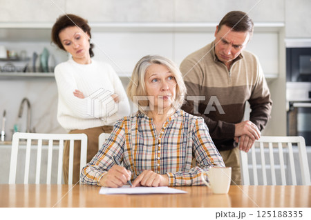 Old woman compiling heritage document while middle-aged man and woman competing in the kitchen 125188335