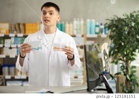 Skilled young man pharmacist standing at prescription counter and offering medicinal products to clients at pharmacy shop Skilled young man pharmacist standing at prescription counter and offering medicinal products to clients at pharmacy shop 125191151