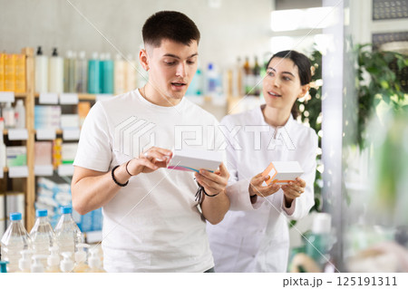 Guy chooses pills in modern pharmacy. Female pharmacist helps with choosing medicine 125191311