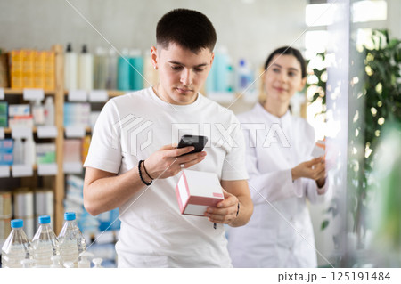 Happy young male customer scan QR code on label and selecting cream or skin care product in drugstore. Female pharmacist helping with choosing and offering variations of medicines 125191484