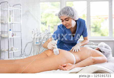 Female cosmetologist performing procedure using cooling attachment on woman client buttocks 125191721