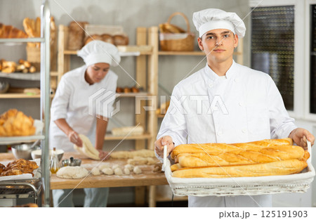 Young male baker holding baguettes in wicker basket Young male baker holding baguettes in wicker basket 125191903