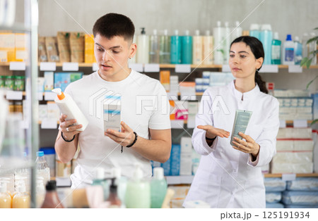 Man chooses skin cream in modern pharmacy. Female pharmacist helps with choosing medicine Man chooses skin cream in modern pharmacy. Female pharmacist helps with choosing medicine 125191934