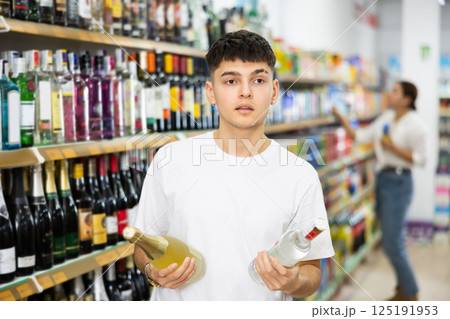 Young male customer deciding on buying bottle of wine or vodka Young male customer deciding on buying bottle of wine or vodka 125191953