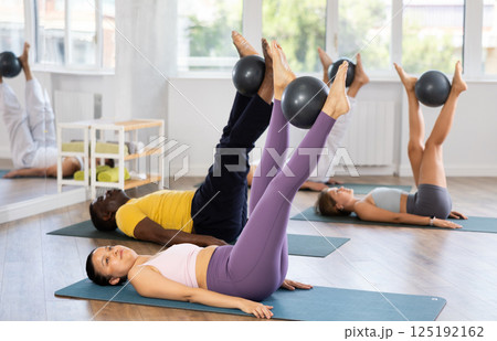 Multinational group of people lie on mats, hold Pilates ball between their knees and perform abdominal exercises during group exercise in fitness studio 125192162