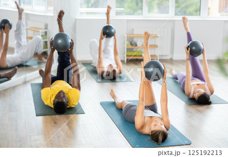 Multinational students repeats movements of coach, lying on mat with hands with fitball up and down Multinational students repeats movements of coach, lying on mat with hands with fitball up and down 125192213