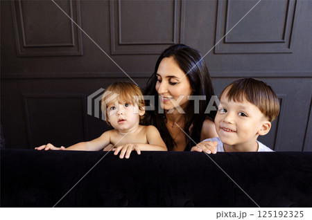 Young mother and her two little boys are playing at home. Woman in casual clothes is having fun with her cute sons in living room. Mom and her kids are laughing and hiding behind couch. 125192325