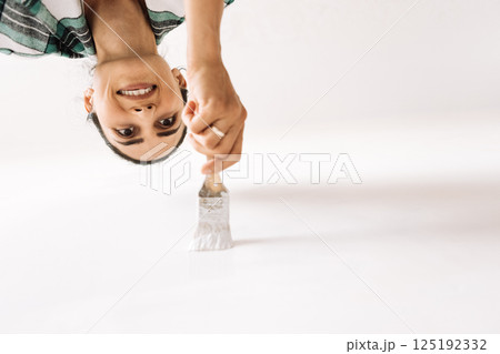 Young woman in casual clothes paints wall in house in white. Bottom view. Girl is holding brush. Smiling painter is working. Female applies paint to inner wall of house. Young woman in casual clothes paints wall in house in white. Bottom view. Girl is holding brush. Smiling painter is working. Female applies paint to inner wall of house. 125192332