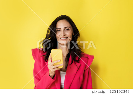 Studio portrait of a surprised young brunette looking at her phone on a bright yellow backgroun 125192345