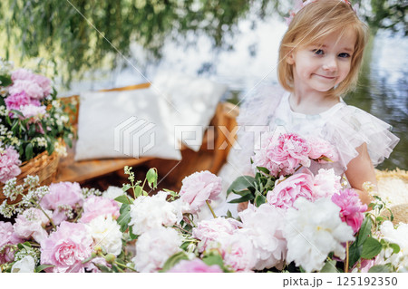 Portrait of a cute little girl in an elegant dress on a wooden boat among pink peonies on the pond or lake 125192350