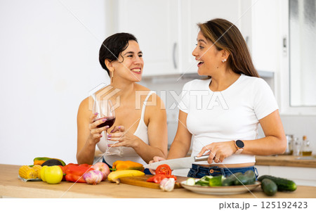 Happy female friends with glasses of red wine chatting and preparing salad together in kitchen 125192423