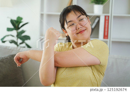 A plump Asian woman is stretching her muscles while sitting on a sofa in her living room at home. 125192674