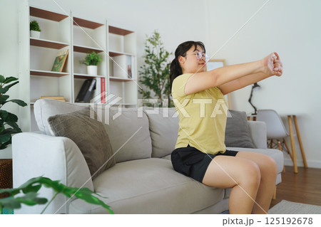 A plump Asian woman is stretching her muscles while sitting on a sofa in her living room at home. 125192678