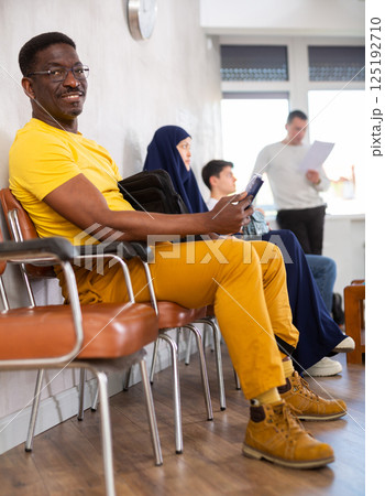 Positive man communicates on mobile phone while sitting on chair in the company office waiting for specialist 125192710