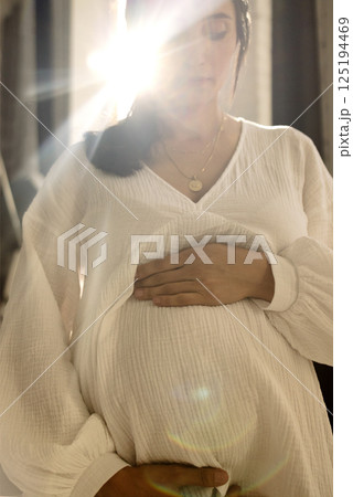 Close-up of a young pregnant woman gently holding her belly. Beautiful natural sunlight. An attractive brunette expecting a baby touches her stomach. Close-up of a young pregnant woman gently holding her belly. Beautiful natural sunlight. An attractive brunette expecting a baby touches her stomach. 125194469