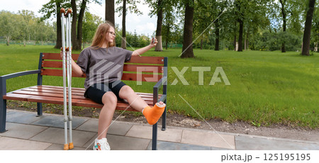 Teenage girl with a cast on her leg seated on a park bench, taking a thoughtful selfie to document the difficulties of her injury and the time she's had to spend in recovery during the summer 125195195