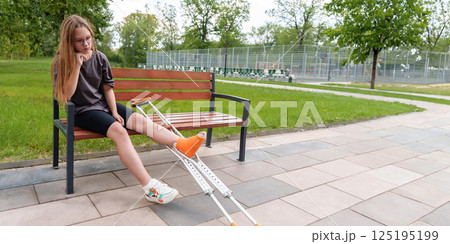 Melancholy teenage girl with a broken leg sits on a park bench, her crutches resting beside her, during a peaceful summer day 125195199