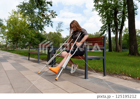 Teen girl struggles to stand from park bench, supporting cast-bound leg on crutches after unexpected injury - healthcare, rehab, disability brands 125195212
