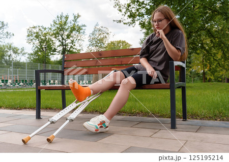Dejected teen girl with cast on leg sits on park bench, conveying injury frustration and limited mobility, healthcare, insurance, youth rehab 125195214