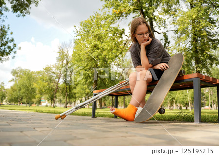 teenage girl, her leg in a cast, sits on a park bench, her gaze fixed on the skateboard that led to her injury, a bittersweet moment of reflection 125195215