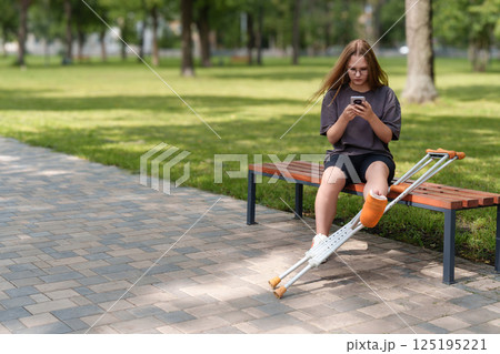 Relaxing on a park bench, a young girl with a bandaged leg uses her phone, her crutches within easy reach in the peaceful 125195221