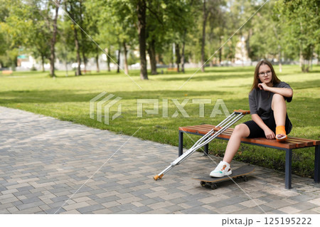 Disheartened girl resting on a park bench with crutches and skateboard nearby 125195222
