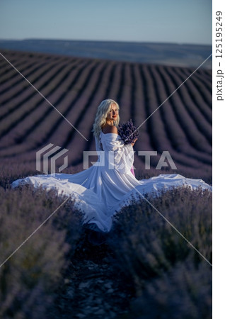 Blonde woman poses in lavender field at sunset. Happy woman in white dress holds lavender bouquet. Aromatherapy concept, lavender oil, photo session in lavender 125195249