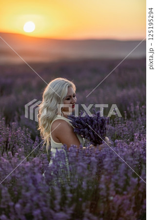 Blonde woman poses in lavender field at sunset. Happy woman in white dress holds lavender bouquet. Aromatherapy concept, lavender oil, photo session in lavender 125195294
