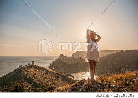 A blonde woman stands on a hill overlooking the ocean. She is wearing a white dress and she is enjoying the view. A blonde woman stands on a hill overlooking the ocean. She is wearing a white dress and she is enjoying the view. 125195308