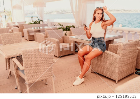 Cafe woman sitting at a table with a cell phone in her hand. She is looking at the screen and she is focused on her phone. The scene takes place in a restaurant or cafe, with multiple chairs. 125195315