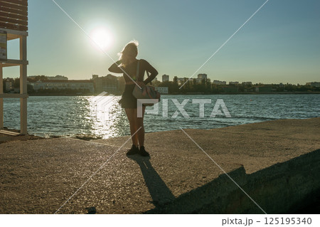 Woman Silhouette Lake Sunset - A lone woman stands on the edge of a lake, her silhouette backlit by the setting sun. The city skyline can be seen in the distance. Woman Silhouette Lake Sunset - A lone woman stands on the edge of a lake, her silhouette backlit by the setting sun. The city skyline can be seen in the distance. 125195340