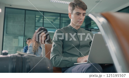 Young handsome man with a suitcase sitting working his laptop at airport terminal hall while waiting landing for her flight, tourist men in international airport, travel trip 125195507