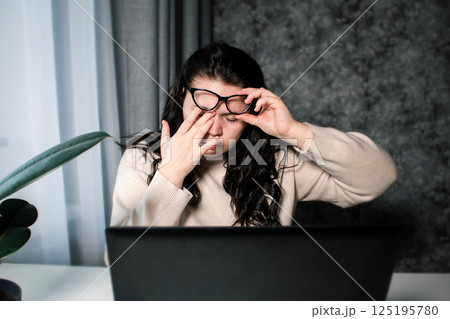 Tired woman working at home with laptop holding eyeglasses rubbing eyes and feeling fatigue. Stress and frustration concept. Tired eyes Tired woman working at home with laptop holding eyeglasses rubbing eyes and feeling fatigue. Stress and frustration concept. Tired eyes 125195780