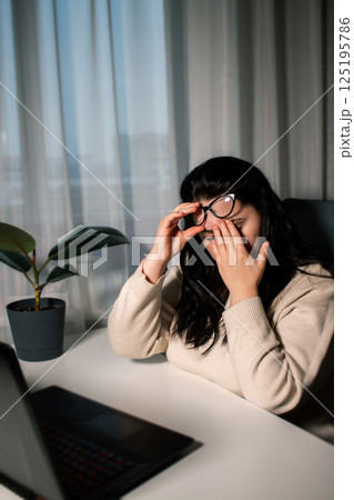 Symptoms of overworking. Young tired female freelancer in eyeglasses sitting in front of computer rubbing eyes feeling eye strain during work Symptoms of overworking. Young tired female freelancer in eyeglasses sitting in front of computer rubbing eyes feeling eye strain during work 125195786