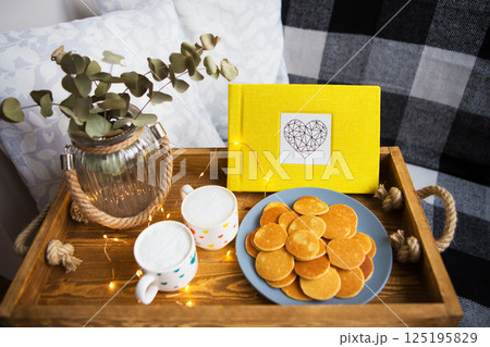 Cozy breakfast in bed scene with mini pancakes, coffee, and a yellow book on a wooden tray 125195829