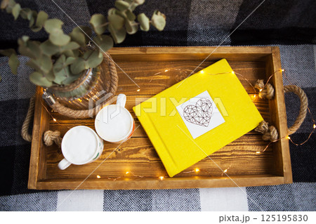Cozy autumn scene with yellow book, mugs, and eucalyptus in a rustic wooden tray, decorated with fairy lights 125195830