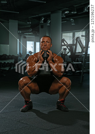 dedicated athlete executes a squat while holding a dumbbell showcasing impressive strength and physique in a well equipped gym. Bright natural light enhances the training atmosphere. 125196237