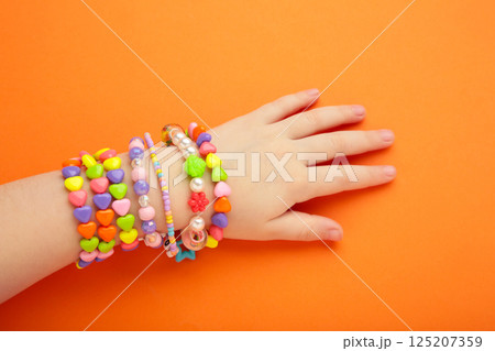 Child wears bracelets on his hands. Kids handmade beaded jewelry on orange background. 125207359