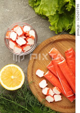 Tasty crab sticks, pieces of lemon and salad on wooden plate on grey background. Tasty crab sticks, pieces of lemon and salad on wooden plate on grey background. 125207746