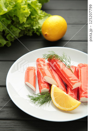 Tasty crab sticks, pieces of lemon and salad on plate on black background. Tasty crab sticks, pieces of lemon and salad on plate on black background. 125207799