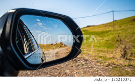 Car side mirror reflecting rural countryside road with blue sky and green hills Car side mirror reflecting rural countryside road with blue sky and green hills 125210373