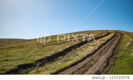 Dirt road winding uphill through green meadow under clear blue sky 125210376