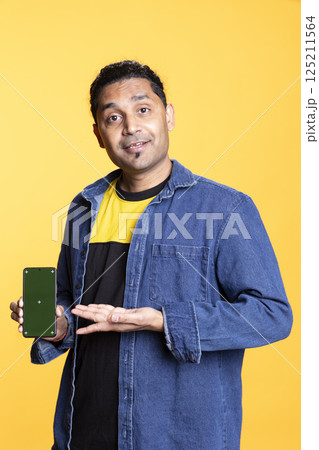 Indian content creator presenting green screen on mobile device, standing against orange background in the studio. Young male model wearing casual clothes and a man bun, internet channel. 125211564