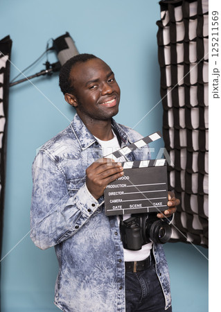 Portrait of smiling african american man standing in studio, holding clapperboard, ready for a shoot. Black male photographer in denim jacket grasping a film slate, standing among lighting equipment. 125211569
