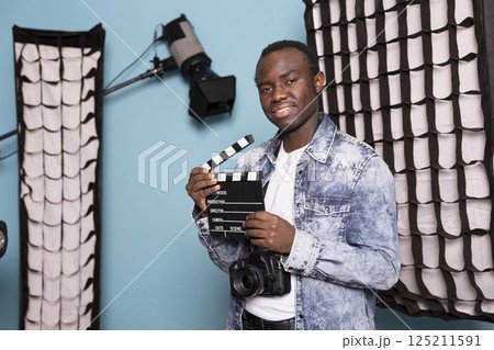 African american filmmaker standing in studio surrounded by lighting equipment, holding a clapperboard. Portrait of black male photographer with camera hanging over his neck and film slate. African american filmmaker standing in studio surrounded by lighting equipment, holding a clapperboard. Portrait of black male photographer with camera hanging over his neck and film slate. 125211591