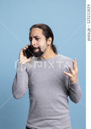 Furious young man getting mad and upset during a phone call with a friend, yelling and feeling angry against blue background. Male adult being irritated and aggressive with his words. 125211624
