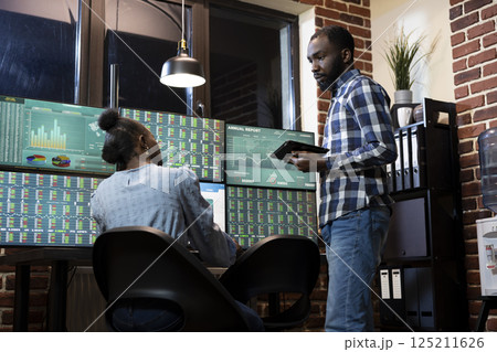 African American forex traders exchange investment strategies while tracking real time stock market transactions during night shift in brick wall office. Teamwork discussion at tech savvy workstation. 125211626