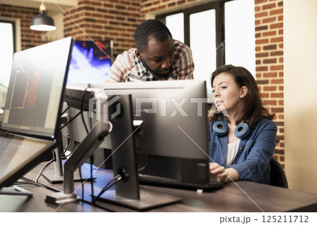 African american man standing and discussing with caucasian woman at desk, analyzing product designs on computer. Diverse employees cooperate in preparing creative projects in brick wall office. African american man standing and discussing with caucasian woman at desk, analyzing product designs on computer. Diverse employees cooperate in preparing creative projects in brick wall office. 125211712